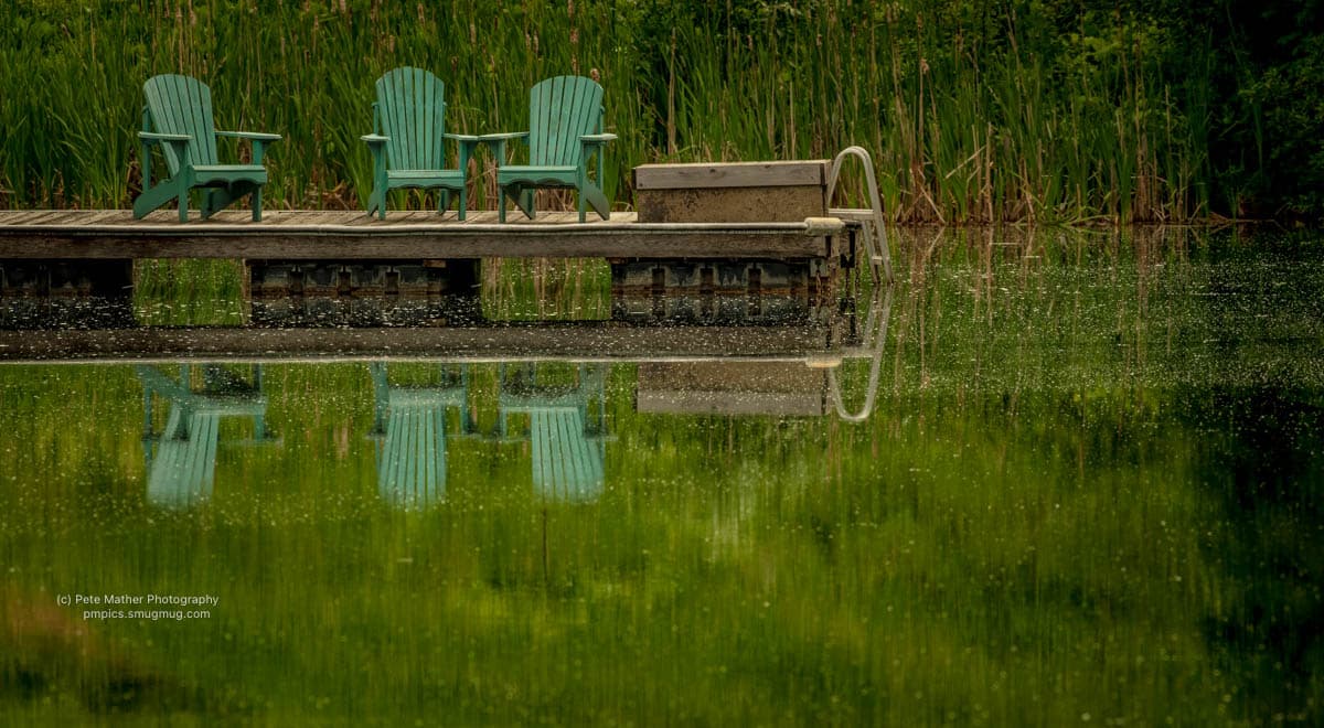 Three teal chairs on a dock reflected in a still pond